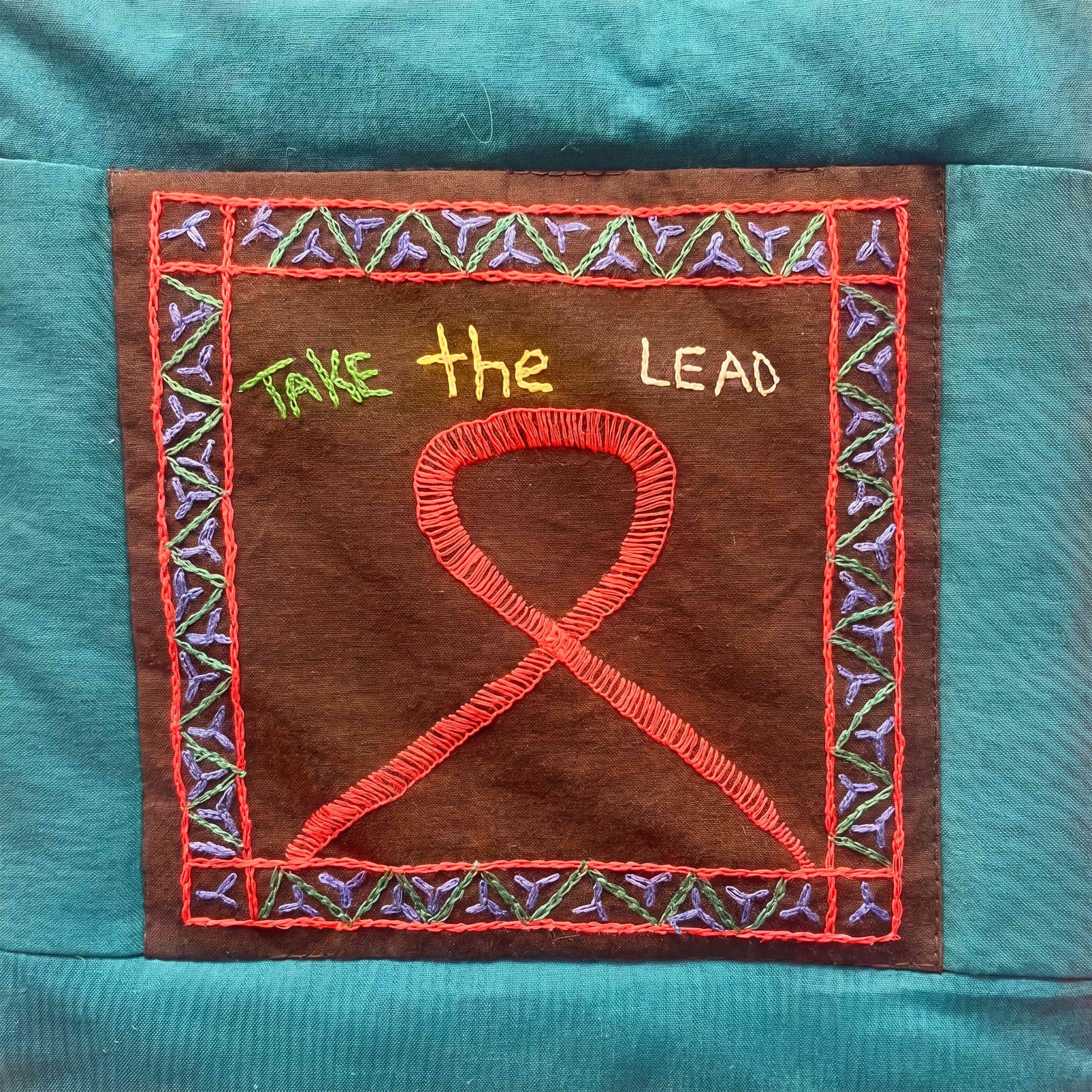 A handmade AIDS quilt panel embroidered with 'Take the Lead' and a red ribbon