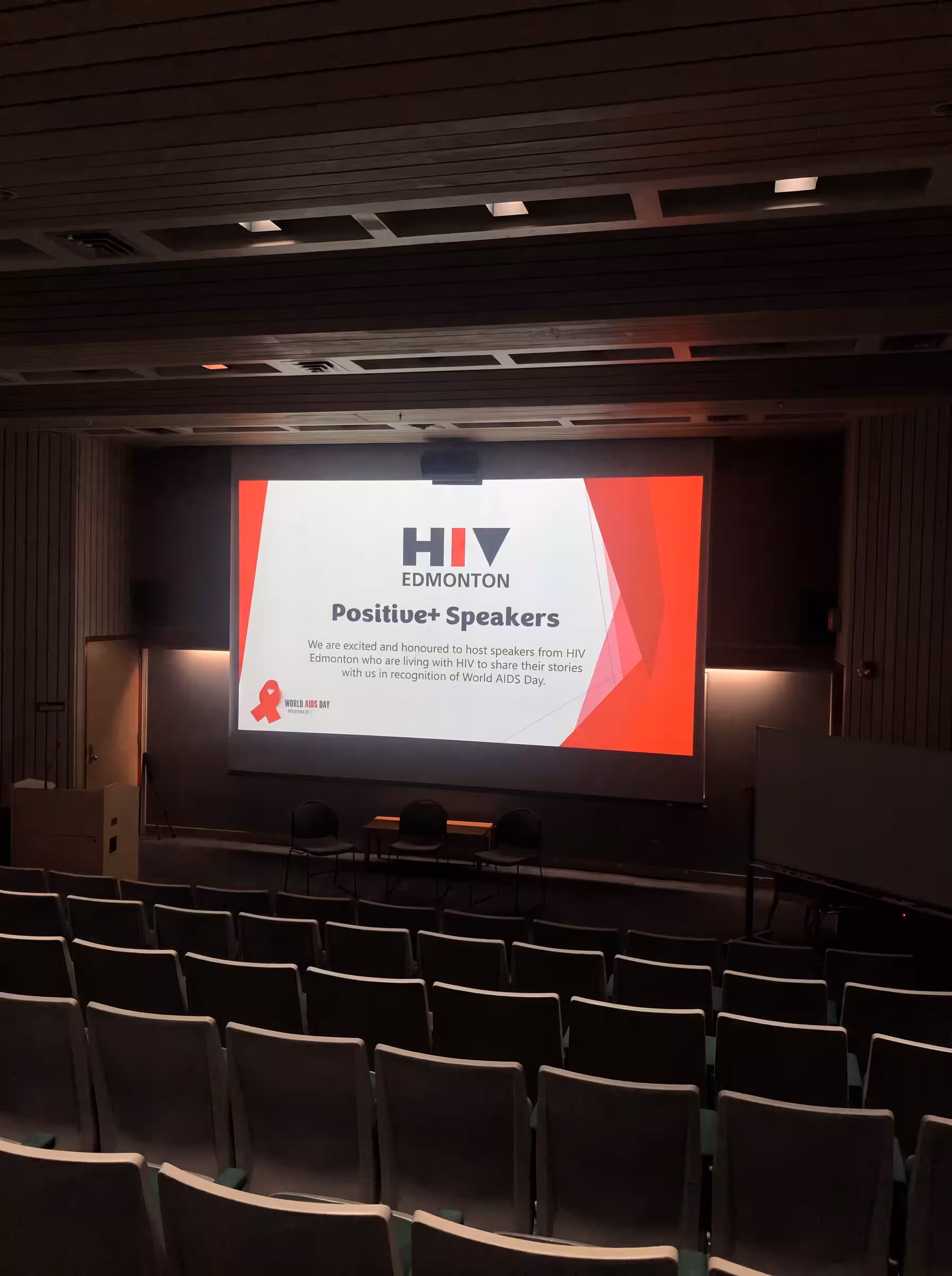 An auditorium with a stage and an HIV Edmonton 'Positive Speakers' presentation slide