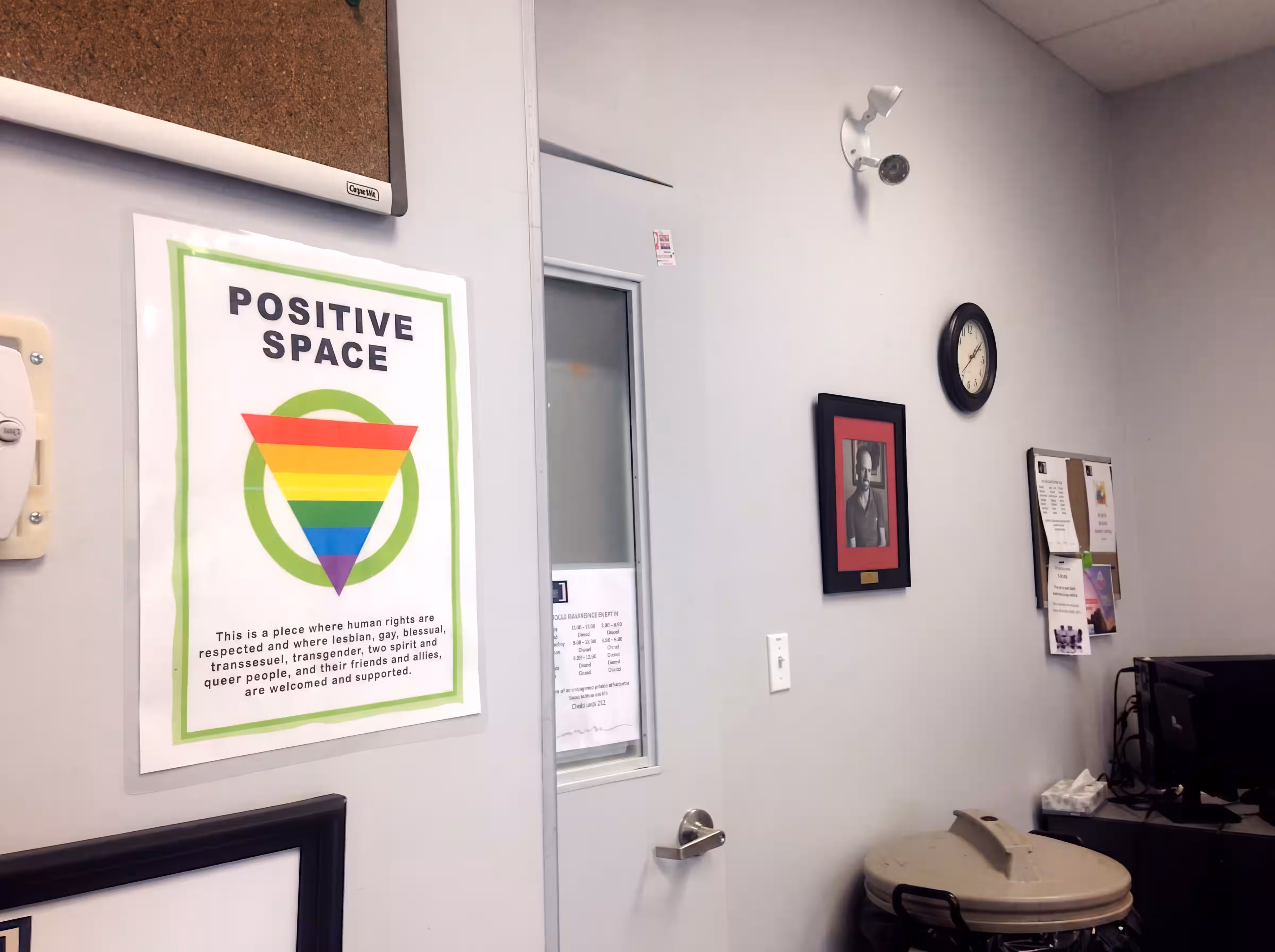 A welcoming room with a rainbow 'Positive Space' poster, bulletin boards, and a clock